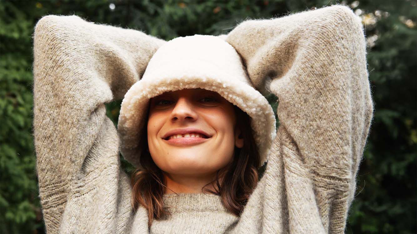 Woman in cozy sweater with hood smiles relaxed and stretches her arms over her head.